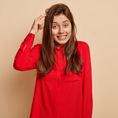 Puzzled unsure delighted young woman scratches head and looks directly at camera, makes decision, recieves good proposal, wears red shirt, poses over light beige background, uses imagination